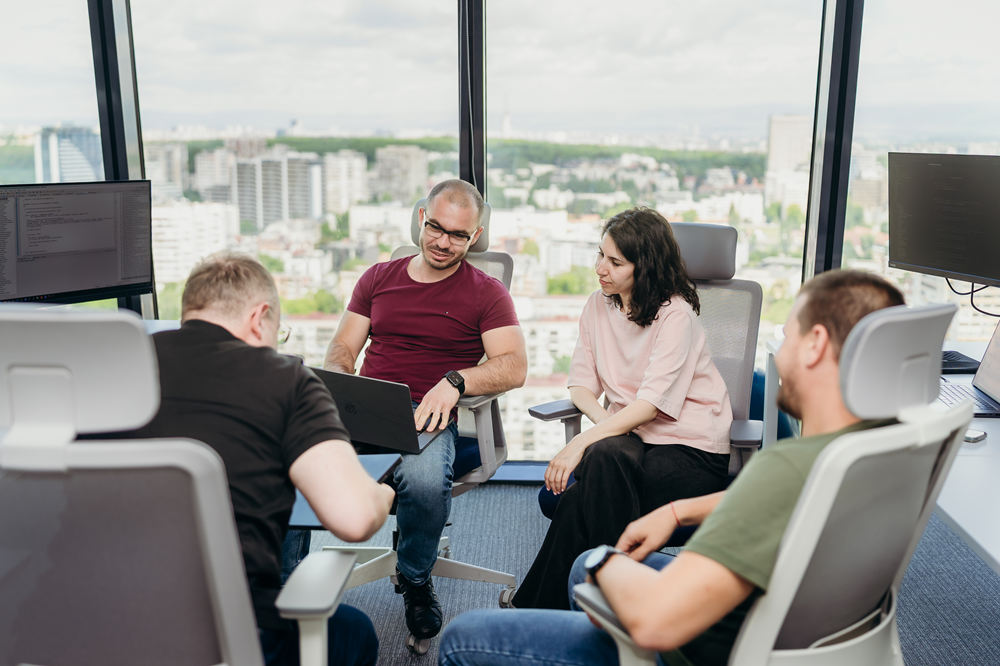 Four coworkers in a glass-walled office discuss a laptop screen with a city skyline in the background, collaborative scene.